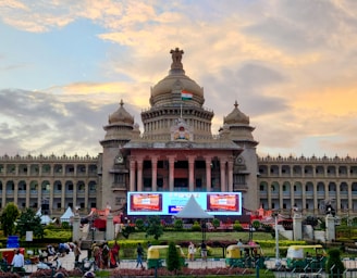 A grand historical building with domed towers and an Indian flag displayed prominently on the top. The structure features intricate architectural details and is flanked by gardens. In the foreground, there are auto rickshaws and pedestrians, while the sky is a vibrant mix of yellows and blues from the setting sun.