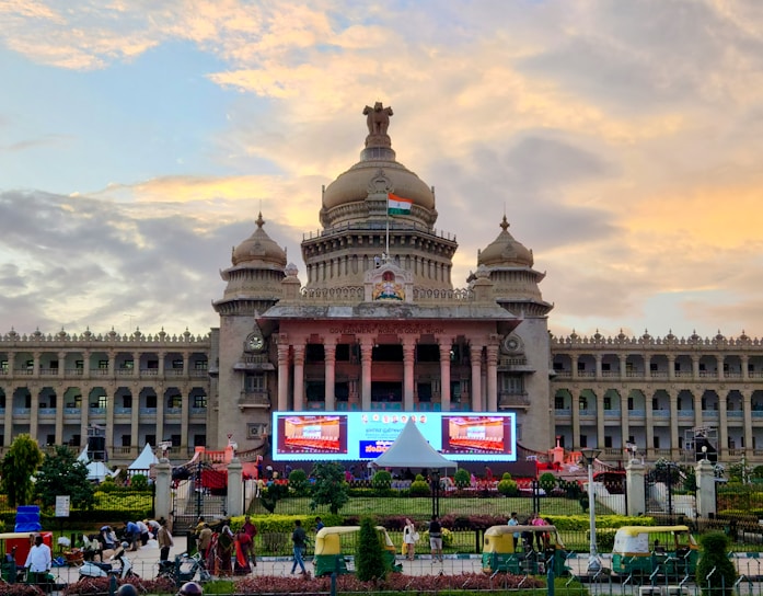 A grand historical building with domed towers and an Indian flag displayed prominently on the top. The structure features intricate architectural details and is flanked by gardens. In the foreground, there are auto rickshaws and pedestrians, while the sky is a vibrant mix of yellows and blues from the setting sun.