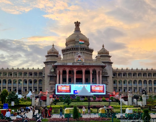 A grand historical building with domed towers and an Indian flag displayed prominently on the top. The structure features intricate architectural details and is flanked by gardens. In the foreground, there are auto rickshaws and pedestrians, while the sky is a vibrant mix of yellows and blues from the setting sun.