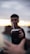A person is offering a traditional mate cup with a metal straw, with their face blurred in the background. The scene appears to take place outdoors during sunset or sunrise, indicated by the soft gradient of light in the sky.