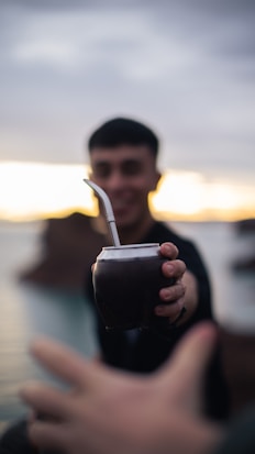 A person is offering a traditional mate cup with a metal straw, with their face blurred in the background. The scene appears to take place outdoors during sunset or sunrise, indicated by the soft gradient of light in the sky.