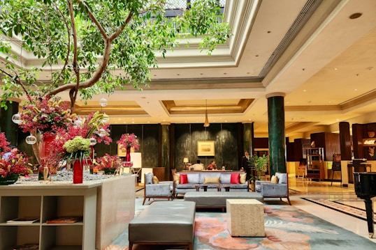 A luxurious hotel lobby with elegant decor, featuring comfortable seating arrangements adorned with colorful pillows. There is a large indoor tree, and vibrant floral arrangements add a touch of color. The space is well-lit, with high ceilings and sophisticated lighting fixtures. Columns and a reception area can be seen in the background.