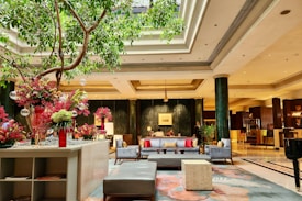 A luxurious hotel lobby with elegant decor, featuring comfortable seating arrangements adorned with colorful pillows. There is a large indoor tree, and vibrant floral arrangements add a touch of color. The space is well-lit, with high ceilings and sophisticated lighting fixtures. Columns and a reception area can be seen in the background.
