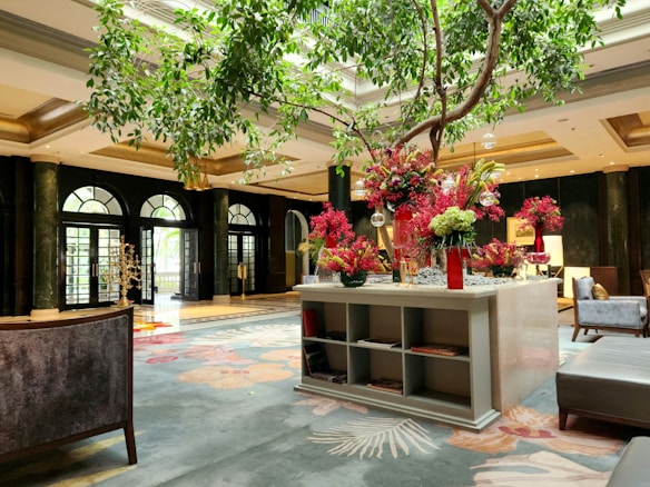 A luxurious hotel lobby with elegant decor, featuring a large floral arrangement with vibrant red flowers on a central table. The area is spacious with high ceilings and large arch windows, letting in natural light. There are comfortable seating areas with plush cushions, and the floor is adorned with an ornate carpet.
