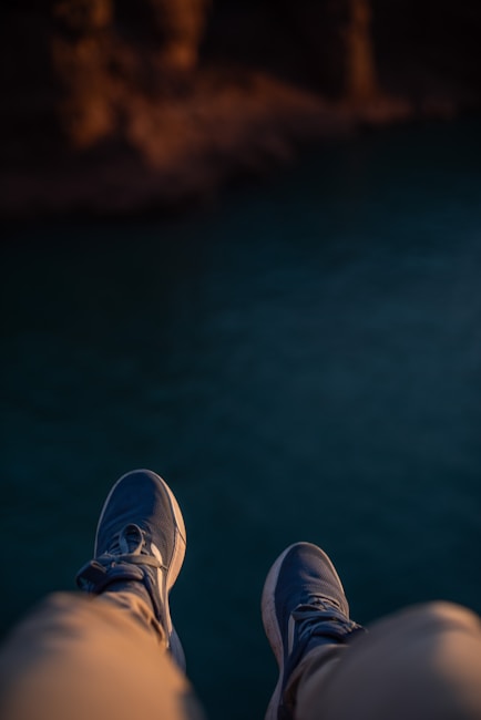 A pair of legs wearing beige pants and blue sneakers dangle over the edge, with a blurred background of water below.