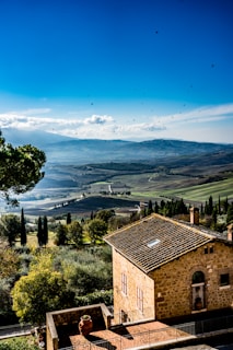 A picturesque rural landscape with rolling hills stretching into the distance, dotted with a few trees and various shades of green and brown fields. In the foreground, an old stone house with a tiled roof stands among dense green foliage. Birds are seen flying in the bright blue sky with a few scattered clouds.