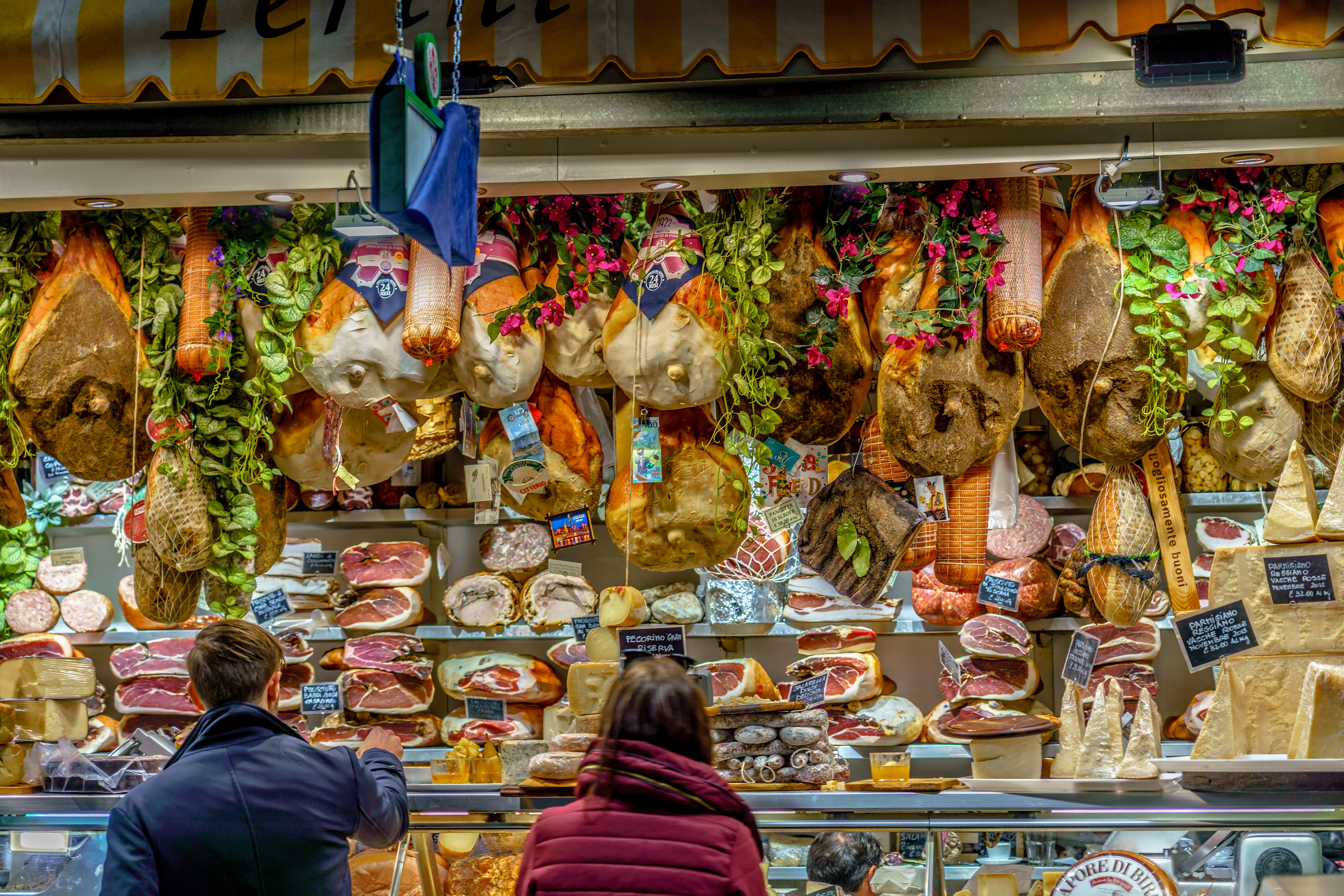 A salumi and cheese counter inside Florence's Mercato Centrale, whole prosciutto legs and sausages hung above a glass case of sliced hams and pecorino, two shoppers waiting to order