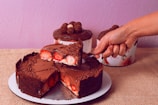 A decadent chocolate cake topped with grated chocolate and filled with fresh strawberries. A hand is slicing a piece of the cake, revealing the layers of chocolate and strawberries. In the background, two more chocolate-topped cakes can be seen, resting on a rustic burlap surface. The wall is painted in a soft lavender shade, adding a contrast to the rich brown of the chocolate.