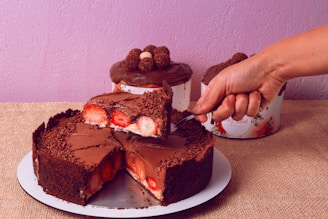 A decadent chocolate cake topped with grated chocolate and filled with fresh strawberries. A hand is slicing a piece of the cake, revealing the layers of chocolate and strawberries. In the background, two more chocolate-topped cakes can be seen, resting on a rustic burlap surface. The wall is painted in a soft lavender shade, adding a contrast to the rich brown of the chocolate.