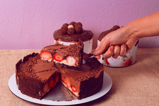 A decadent chocolate cake topped with grated chocolate and filled with fresh strawberries. A hand is slicing a piece of the cake, revealing the layers of chocolate and strawberries. In the background, two more chocolate-topped cakes can be seen, resting on a rustic burlap surface. The wall is painted in a soft lavender shade, adding a contrast to the rich brown of the chocolate.