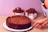 A chocolate cake with a fluffy texture sits on a white plate, partially cut into slices. Behind it, two smaller cakes topped with chocolate frosting and garnished with chocolate truffles are displayed. The background is a light pink wall, and the scene is set on a burlap surface. A hand is holding a utensil, poised to serve the cake.