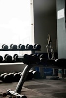An exercise bench is positioned in the foreground next to a large window, with shelves of dumbbells visible in the background. The room is dimly lit, creating a contrast between the bright window light and the dark interior. A piece of gym equipment is also visible on the right side.