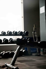 An exercise bench is positioned in the foreground next to a large window, with shelves of dumbbells visible in the background. The room is dimly lit, creating a contrast between the bright window light and the dark interior. A piece of gym equipment is also visible on the right side.