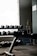 An exercise bench is positioned in the foreground next to a large window, with shelves of dumbbells visible in the background. The room is dimly lit, creating a contrast between the bright window light and the dark interior. A piece of gym equipment is also visible on the right side.