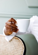 A hand is elegantly holding a white cup against a neutral, muted background. The edge of a woven chair is partially visible, suggesting a relaxed setting.