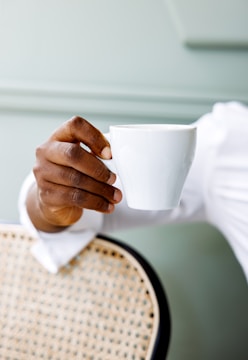 A hand is elegantly holding a white cup against a neutral, muted background. The edge of a woven chair is partially visible, suggesting a relaxed setting.