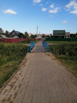 A small footbridge with blue railings crosses over a narrow stream. The pathway is paved with bricks and leads into a rural area with houses in view, surrounded by green fields and patches of trees. The sky is mostly clear with a few scattered clouds.
