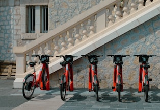 Several red rental bicycles are parked in a row against a stone wall with ornate staircase and balustrade. The setting appears to be in a quiet, urban area with architectural features typical of a historical or European environment.