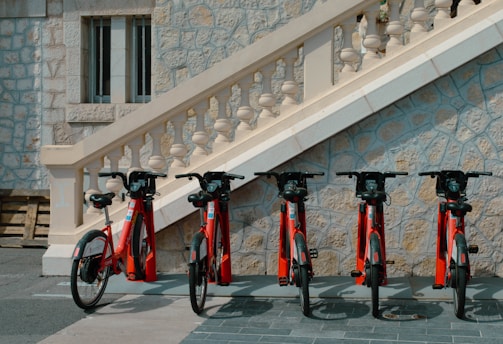Several red rental bicycles are parked in a row against a stone wall with ornate staircase and balustrade. The setting appears to be in a quiet, urban area with architectural features typical of a historical or European environment.