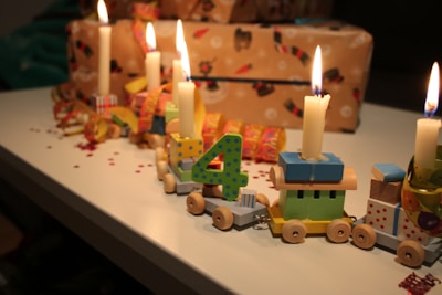 A decorative wooden train with numbered blocks and small lit candles is placed on a table. The train is accompanied by wrapped presents in festive paper. The setting is likely for a birthday celebration, featuring bright colors and small confetti on the surface.