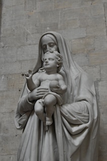 A stone sculpture depicting a motherly figure holding an infant. The mother is draped in robes, and both figures convey a serene expression. The sculpture is set against a muted stone wall background.