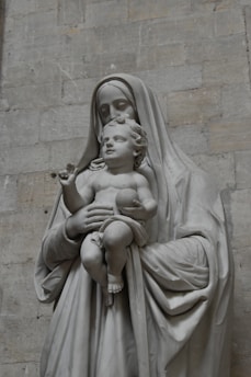 A stone sculpture depicting a motherly figure holding an infant. The mother is draped in robes, and both figures convey a serene expression. The sculpture is set against a muted stone wall background.