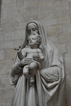 A stone sculpture depicting a motherly figure holding an infant. The mother is draped in robes, and both figures convey a serene expression. The sculpture is set against a muted stone wall background.