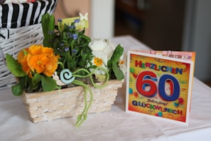 A wicker basket containing a vibrant arrangement of flowers, including orange and white blooms, accompanied by green foliage and a small decorative snail figure. Beside the basket, there is a colorful greeting card with bold text celebrating a 60th birthday in German.