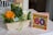 A wicker basket containing a vibrant arrangement of flowers, including orange and white blooms, accompanied by green foliage and a small decorative snail figure. Beside the basket, there is a colorful greeting card with bold text celebrating a 60th birthday in German.