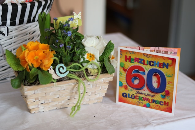 A wicker basket containing a vibrant arrangement of flowers, including orange and white blooms, accompanied by green foliage and a small decorative snail figure. Beside the basket, there is a colorful greeting card with bold text celebrating a 60th birthday in German.