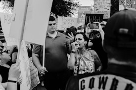 A black and white photograph of a protest scene with people holding signs and megaphones. The central figure is passionately speaking into a microphone, surrounded by others capturing the moment on their phones.