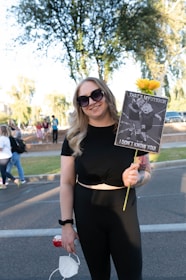 A person wearing sunglasses and a black outfit is holding a sign with a humorous message about a uterus, accompanied by a yellow flower. There are multiple people in the background on a street lined with trees.