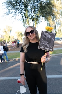 A person wearing sunglasses and a black outfit is holding a sign with a humorous message about a uterus, accompanied by a yellow flower. There are multiple people in the background on a street lined with trees.