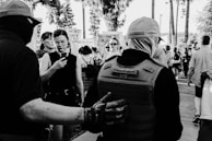 A group of people gathered in a protest setting, some holding signs with messages. One person in the foreground wears protective gear, and another is looking at a smartphone. The scene appears to take place outdoors, with trees and a pavement visible.