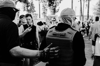 A group of people gathered in a protest setting, some holding signs with messages. One person in the foreground wears protective gear, and another is looking at a smartphone. The scene appears to take place outdoors, with trees and a pavement visible.
