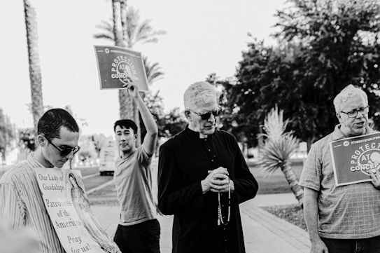 A group of people are outdoors, with two individuals holding signs that read 'Protection at Conception.' One person is dressed in religious attire, holding a rosary, which suggests a prayer or protest setting. The background features palm trees and a pathway, indicating a warm, possibly tropical area.