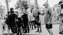 A black and white image depicting a group of people gathered in a public setting with placards. Some individuals are interacting face to face, and one person is speaking through a megaphone. The scene includes palm trees and a sidewalk, suggesting an outdoor location, possibly a protest or demonstration.