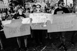 A group of people in a demonstration hold various placards with slogans advocating for women's rights and criticizing patriarchy. The scene is intense and crowded with participants showing strong expressions of activism.