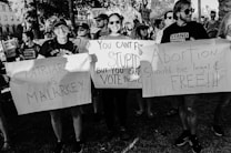 A group of people in a demonstration hold various placards with slogans advocating for women's rights and criticizing patriarchy. The scene is intense and crowded with participants showing strong expressions of activism.