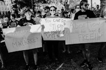 A group of people in a demonstration hold various placards with slogans advocating for women's rights and criticizing patriarchy. The scene is intense and crowded with participants showing strong expressions of activism.