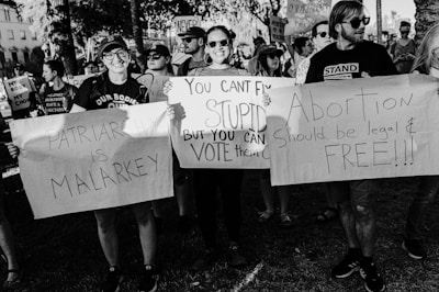 A group of people in a demonstration hold various placards with slogans advocating for women's rights and criticizing patriarchy. The scene is intense and crowded with participants showing strong expressions of activism.
