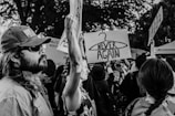 A group of people participating in a protest or rally, holding up signs. One prominent sign depicts a coat hanger with the words 'NEVER AGAIN'. The scene is captured in black and white, giving it a documentary feel. Individuals appear engaged and determined, with a range of expressions. Trees are visible in the background.