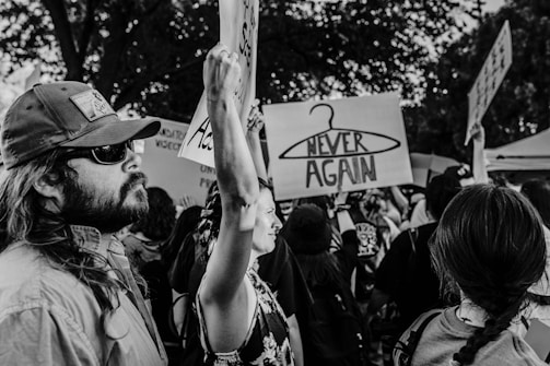 A group of people participating in a protest or rally, holding up signs. One prominent sign depicts a coat hanger with the words 'NEVER AGAIN'. The scene is captured in black and white, giving it a documentary feel. Individuals appear engaged and determined, with a range of expressions. Trees are visible in the background.