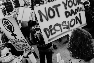 A monochrome image of a crowd at a protest. People are holding signs with bold statements. One sign displays a feminist symbol and the words 'Women's Rights'. Another sign reads 'Not Your Damn Decision'. Various individuals wear expressions of determination and engagement.