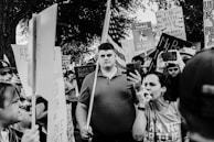 A crowd of people is gathered, holding various signs at what appears to be a protest. Many signs have text supporting or opposing abortion rights. A woman in the foreground speaks through a megaphone, while others hold up phones, likely recording or taking photos. An American flag is partially visible held by one of the participants in the background.