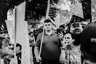 A crowd of people is gathered, holding various signs at what appears to be a protest. Many signs have text supporting or opposing abortion rights. A woman in the foreground speaks through a megaphone, while others hold up phones, likely recording or taking photos. An American flag is partially visible held by one of the participants in the background.
