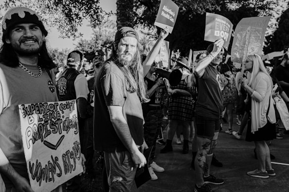A group of people are gathered in a protest, holding signs related to women's rights and freedom of choice. The crowd is diverse, with several individuals prominently featured, one of whom is holding a sign that reads 'Superior Court be like: Rapist Rights Women's Rights.' Others hold signs with messages about healthcare and bodily autonomy. The setting appears to be outdoors on a bright day under the shade of trees.