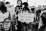 A black and white photograph of a group of people participating in a protest. A young person in the foreground holds a sign with a political statement. Others in the background are also holding signs and appear to be engaged in conversation. The setting is outdoors, possibly in a park or public area.
