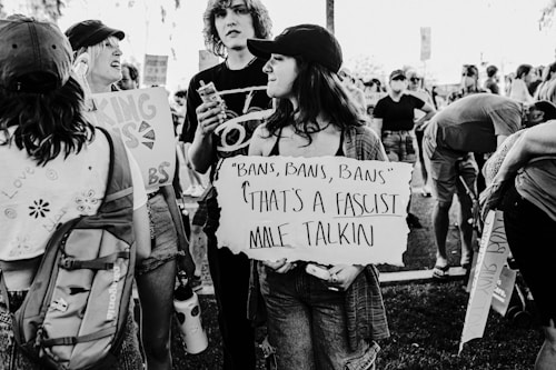 A black and white photograph of a group of people participating in a protest. A young person in the foreground holds a sign with a political statement. Others in the background are also holding signs and appear to be engaged in conversation. The setting is outdoors, possibly in a park or public area.