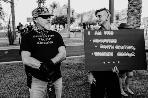 Two people are standing outdoors in a park, engaging in conversation. One person wearing a shirt with the text 'Bikers for Trump Arizona' appears to be a biker with sunglasses, a beard, and a camouflage cap. The other person is holding a sign that reads 'I am pro adoption, birth control, women's choice.' Trees and more people are visible in the background.